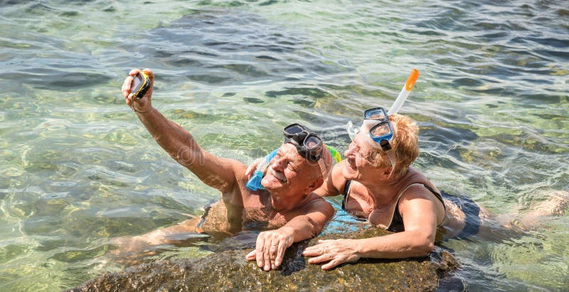 Pares aposentados felizes que tomam o selfie na excursão tropical do mar com câmera da água e a máscara do tubo de respiração - v