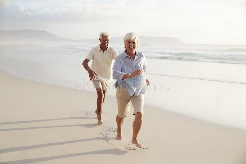 Pares aposentados sênior que correm ao longo da praia do verão junto foto de stock Pares aposentados sênior que correm ao longo da praia do verão junto foto de stock