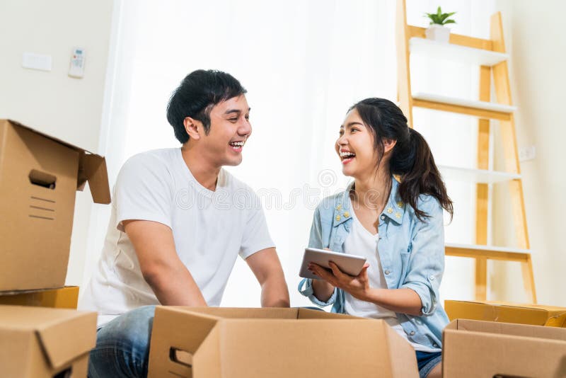 Pares asiáticos novos felizes que movem-se dentro para a casa nova, usando coisas de organização da tabuleta digital e desembalan imagem de stock royalty free
