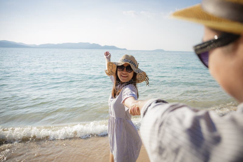 Pares asi?ticos que apreciam f?rias da praia na praia, amiga de sorriso que anda com m?o da terra arrendada de seu noivo na praia fotografia de stock Pares asi?ticos que apreciam f?rias da praia na praia, amiga de sorriso que anda com m?o da terra arrendada de seu noivo na praia fotografia de stock