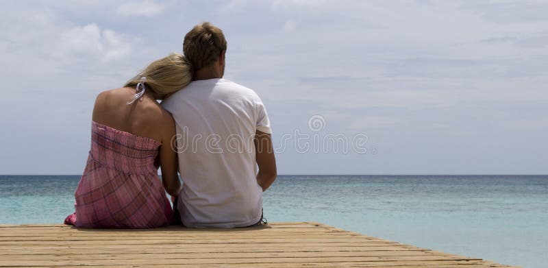 A young couple sits on a dock looking out over the calm blue waters of the Caribbean. Shot in the Dominican Republic at Bahia de las Aguilas. A young couple sits on a dock looking out over the calm blue waters of the Caribbean. Shot in the Dominican Republic at Bahia de las Aguilas.