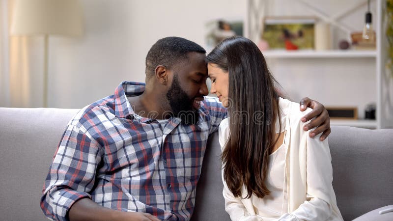 Pares de amor de la raza mixta que tocan las frentes y que abrazan en el sofá, dulzura imagen de archivo Pares de amor de la raza mixta que tocan las frentes y que abrazan en el sofá, dulzura imagen de archivo