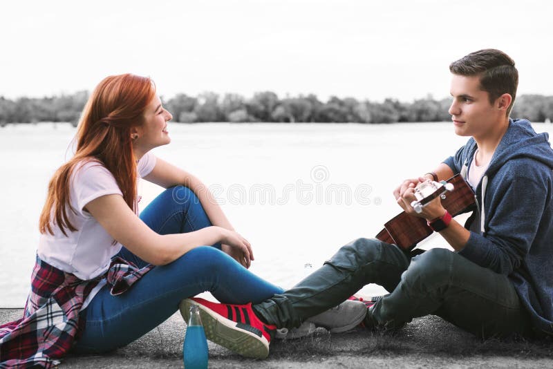 Pares de los adolescentes de emisión felices que disfrutan de la tarde agradable con la guitarra cerca del río fotos de archivo libres de regalías Pares de los adolescentes de emisión felices que disfrutan de la tarde agradable con la guitarra cerca del río fotos de archivo libres de regalías