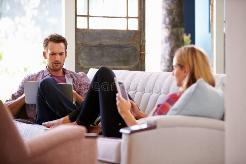 Pares em casa em dispositivos de Sofa In Lounge Using Digital fotografia de stock Pares em casa em dispositivos de Sofa In Lounge Using Digital fotografia de stock