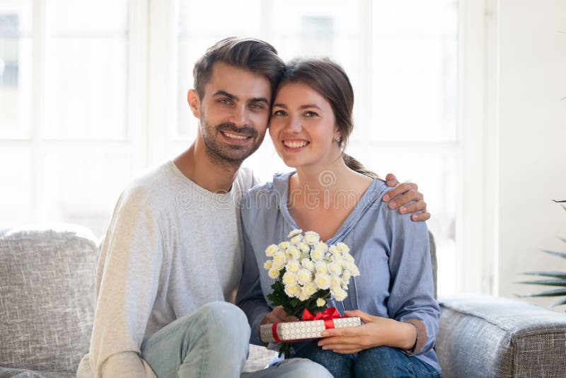 Pares felices que se sientan en el sofá con las flores y la caja de regalo fotos de archivo Pares felices que se sientan en el sofá con las flores y la caja de regalo fotos de archivo