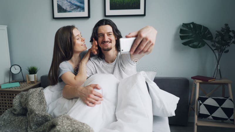 Pares felices que toman el selfie en cama usando la presentación sonriente de la cámara del smartphone