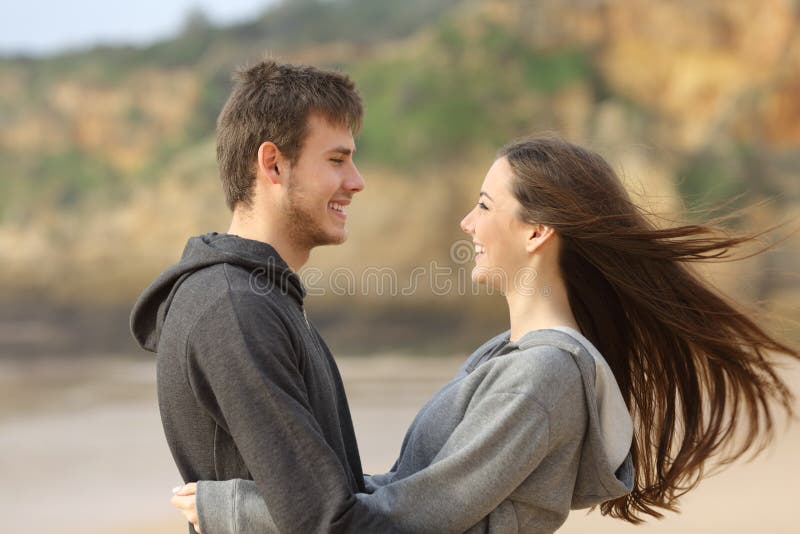 Side view of happy teenager couple dating and flirting hugging and facing on the beach while the wind moves her hair. Side view of happy teenager couple dating and flirting hugging and facing on the beach while the wind moves her hair