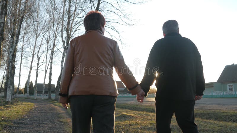 Pares idosos loving que andam no parque no por do sol Próximo há uma estrada Uma mulher em um casaco de cabedal marrom O homem