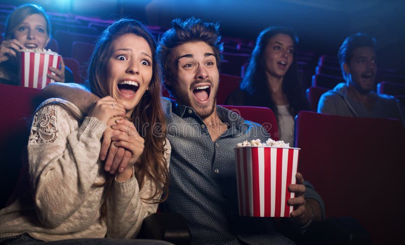 Young scared couple at the cinema watching an horror movie and screaming, she is holding her boyfriend's hand. Young scared couple at the cinema watching an horror movie and screaming, she is holding her boyfriend's hand