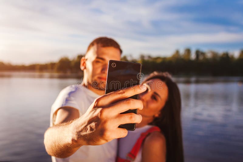 Pares jovenes felices que toman el selfie en la orilla del río en la puesta del sol Mujer y hombre que se divierten Primer del sm imagen de archivo Pares jovenes felices que toman el selfie en la orilla del río en la puesta del sol Mujer y hombre que se divierten Primer del sm imagen de archivo