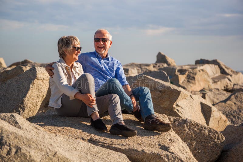 Pares mayores felices que se sientan en rocas por el mar imagen de archivo libre de regalías Pares mayores felices que se sientan en rocas por el mar imagen de archivo libre de regalías