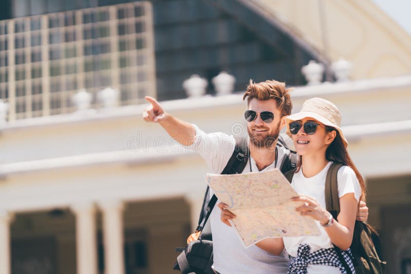 Pares multiétnicos del viajero usando mapa local genérico junto el día soleado Viaje de la luna de miel, turista del backpacker