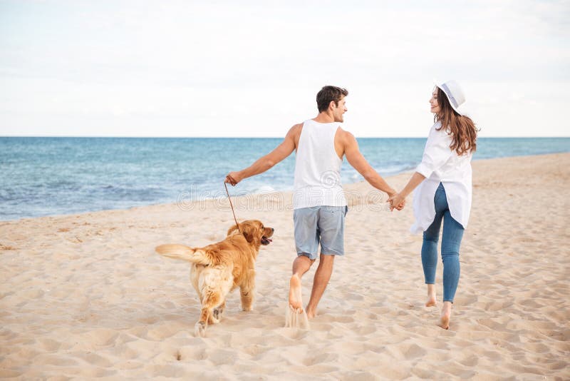 Pares novos alegres felizes que correm na praia com seu cão