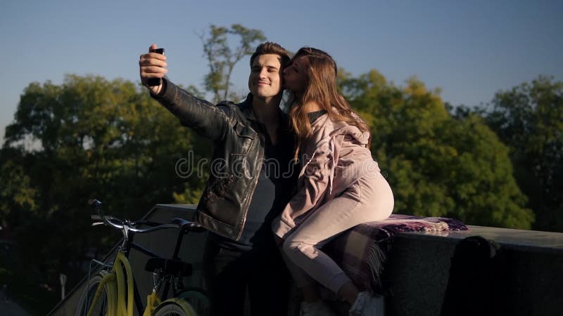 Pares novos bonitos que tomam o selfie pelo telefone celular na rua quando menina que senta-se no parapeito Pares bonitos