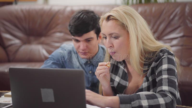 Pares novos do retrato que sentam-se na frente de um caderno em casa Homem moreno considerável e mulher loura bonito que trabalha