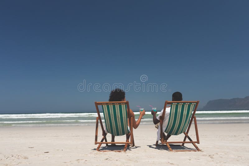 Pares novos que relaxam no vadio do sol na praia no dia ensolarado