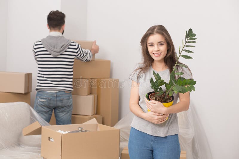 Young couple relocating to new apartment and unpacking boxes. Front view of young women holding plant and men standing behind unpacking boxes. Young couple relocating to new apartment and unpacking boxes. Front view of young women holding plant and men standing behind unpacking boxes.