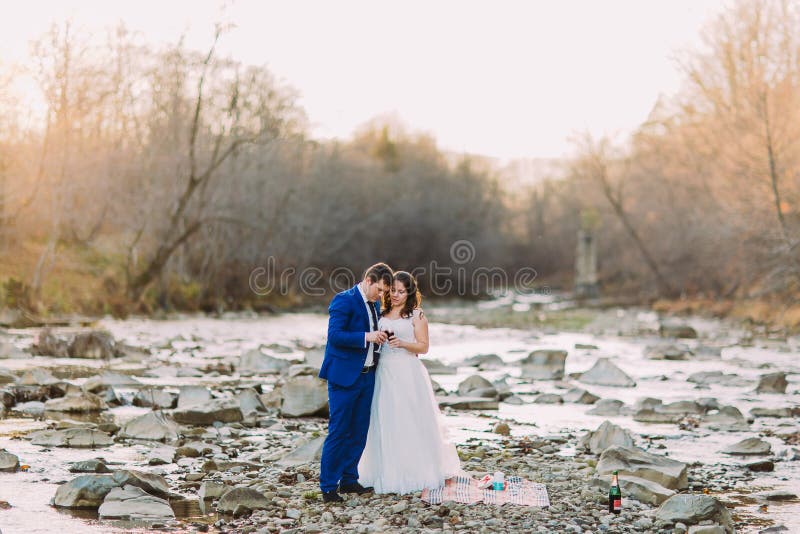 Pares nupciais novos românticos do vinho bebendo no banco de rio rochoso do seixo com Forest Hills e córrego imagem de stock Pares nupciais novos românticos do vinho bebendo no banco de rio rochoso do seixo com Forest Hills e córrego imagem de stock
