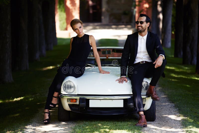 Pares románticos y de moda que presentan en el coche de lujo del cabriolé al aire libre en verano fotos de archivo Pares románticos y de moda que presentan en el coche de lujo del cabriolé al aire libre en verano fotos de archivo