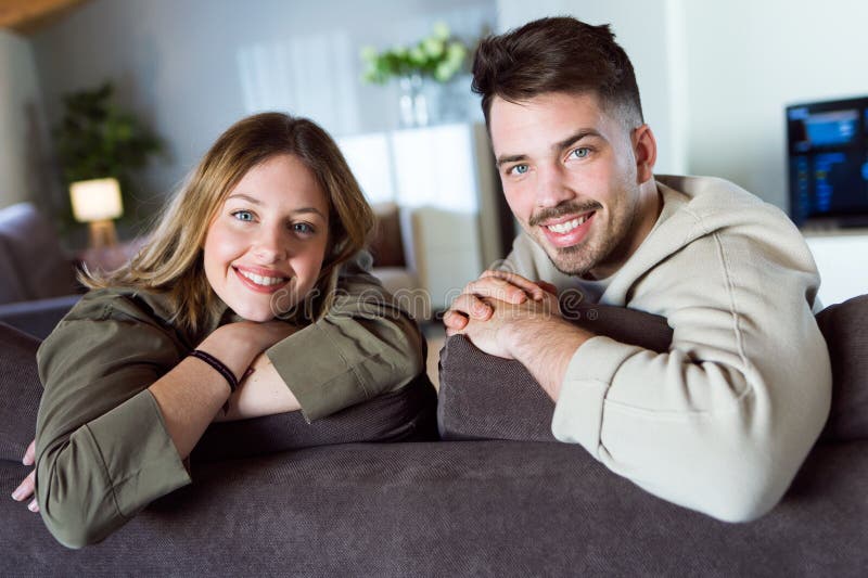 Pares sonrientes jovenes hermosos que miran la cámara y que se sientan en el sofá en casa fotografía de archivo Pares sonrientes jovenes hermosos que miran la cámara y que se sientan en el sofá en casa fotografía de archivo