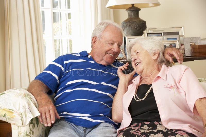 Pares superiores aposentados que sentam-se na casa de Sofa Talking On Phone At junto fotografia de stock royalty free Pares superiores aposentados que sentam-se na casa de Sofa Talking On Phone At junto fotografia de stock royalty free