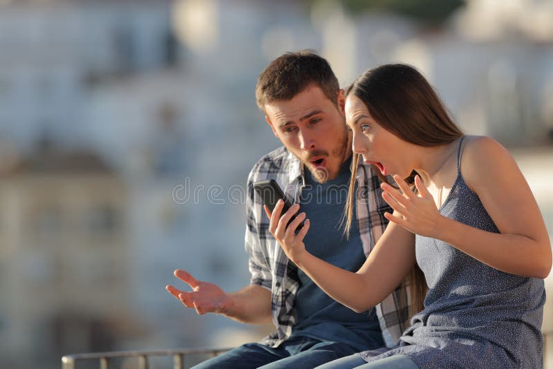 Pares surpreendidos que verificam o telefone esperto em uma cidade imagem de stock Pares surpreendidos que verificam o telefone esperto em uma cidade imagem de stock