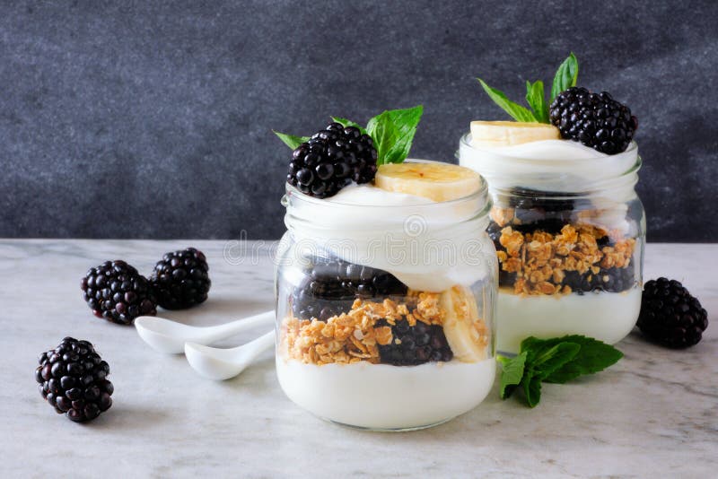 Healthy blackberry and banana yogurt parfaits in mason jars. Still life on dark stone background. Healthy blackberry and banana yogurt parfaits in mason jars. Still life on dark stone background