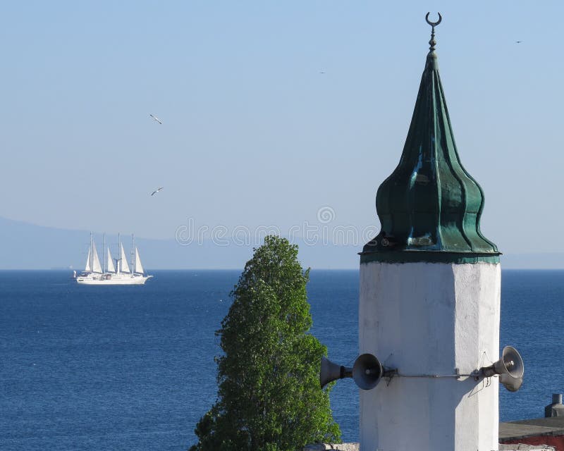 A parte superior do minarete e a planta contra o contexto do mar, o céu e um passado branco da navigação do veleiro imagem de stock A parte superior do minarete e a planta contra o contexto do mar, o céu e um passado branco da navigação do veleiro imagem de stock