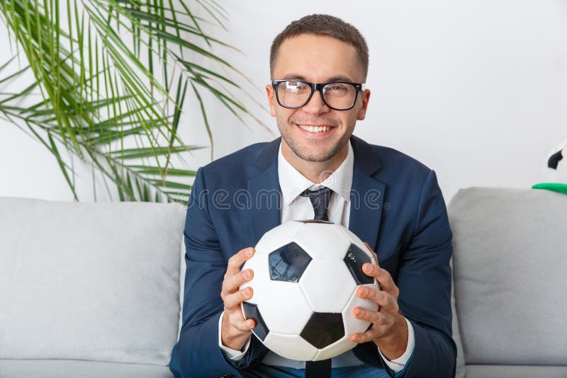 Partita di sorveglianza del giovane tifoso dell'uomo d'affari che guarda sorridere della macchina fotografica fotografia stock Partita di sorveglianza del giovane tifoso dell'uomo d'affari che guarda sorridere della macchina fotografica fotografia stock