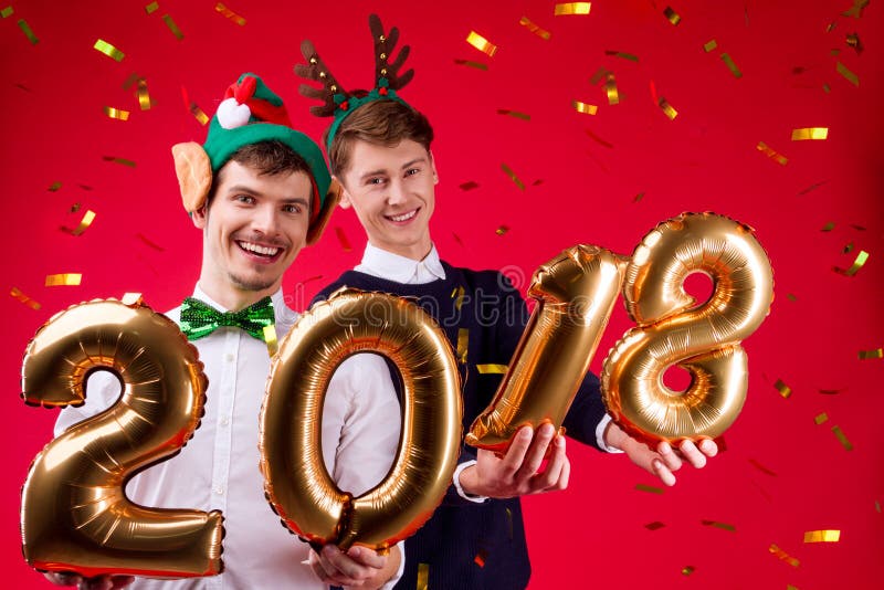 Pair of males / men in decorated new year`s hats: elf and deer horn, holding gold balloons numbers 2018 as symbol of New Year on red background with gold confetti. Pair of males / men in decorated new year`s hats: elf and deer horn, holding gold balloons numbers 2018 as symbol of New Year on red background with gold confetti