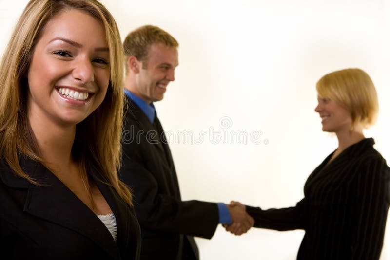 Partnership. Smiling attractive business woman in focus with two business people shaking hands in the background out of focus