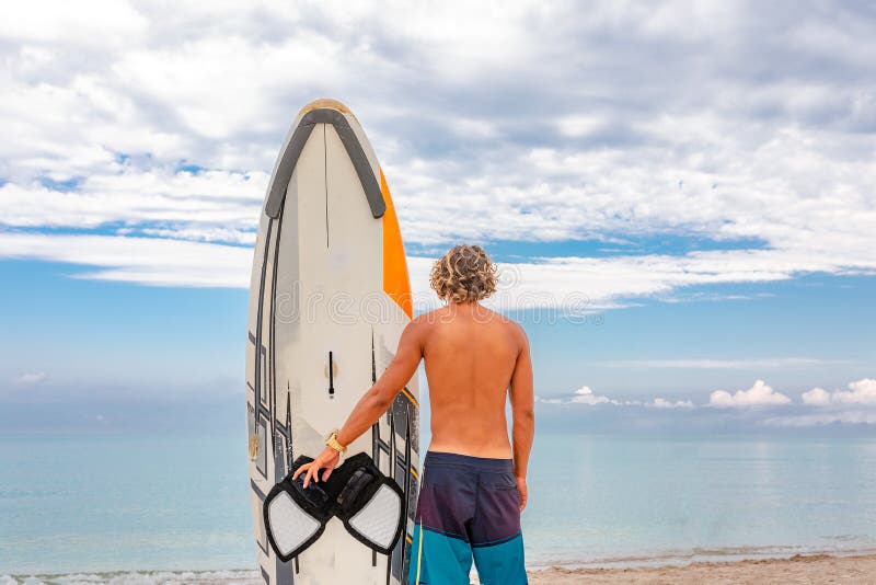 Paseo hermoso del hombre con la espera en blanco blanca del tablero que practica surf para que onda practique surf el punto en la foto de archivo libre de regalías Paseo hermoso del hombre con la espera en blanco blanca del tablero que practica surf para que onda practique surf el punto en la foto de archivo libre de regalías