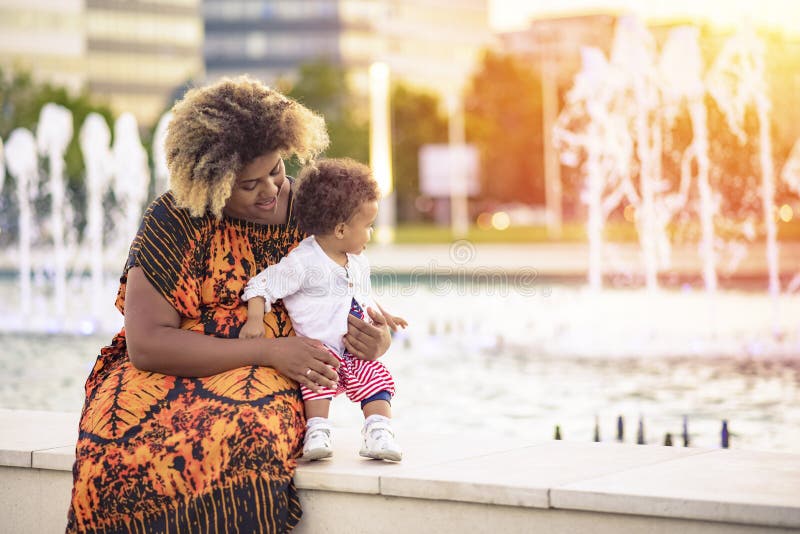 Pasos del ` s primer de la hija del bebé. Madre afroamericana joven que enseña su hija del bebé a cómo caminar cerca de una fuente durante una puesta del sol maravillosa