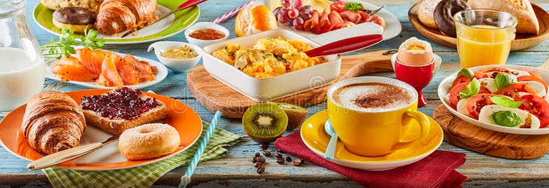 Colorful breakfast spread in a panoramic banner with smoked salmon, Caprese salad, scrambled and boiled egg, meat cheese donut, toast and jam, orange juice and coffee viewed low angle. Colorful breakfast spread in a panoramic banner with smoked salmon, Caprese salad, scrambled and boiled egg, meat cheese donut, toast and jam, orange juice and coffee viewed low angle