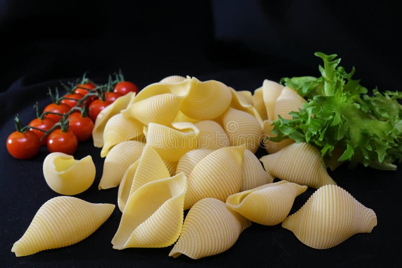 Pasta in the form of shells in a round wooden bowl with cherry tomatoes on a black background. Culinary background stock photo Pasta in the form of shells in a round wooden bowl with cherry tomatoes on a black background. Culinary background stock photo
