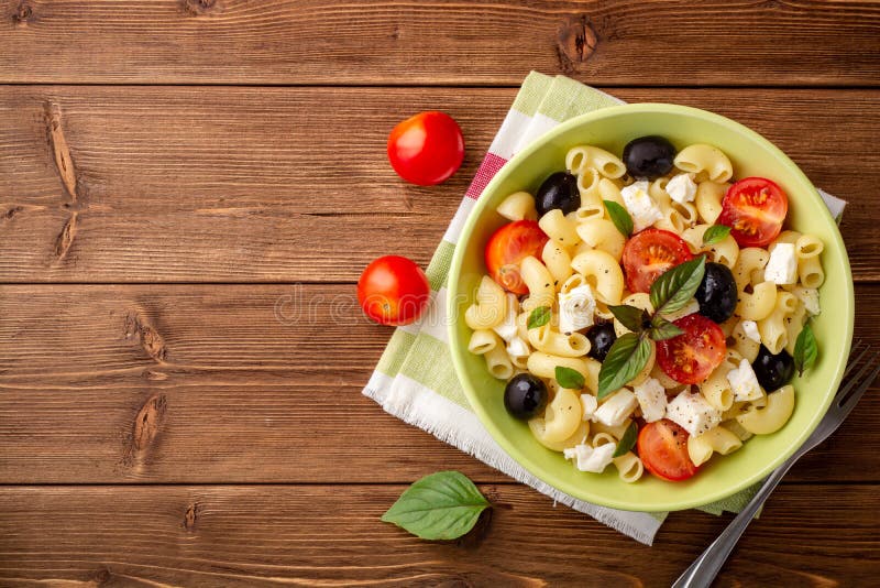 Pasta salad with cherry tomatoes, black olives, feta cheese and basil on wooden background