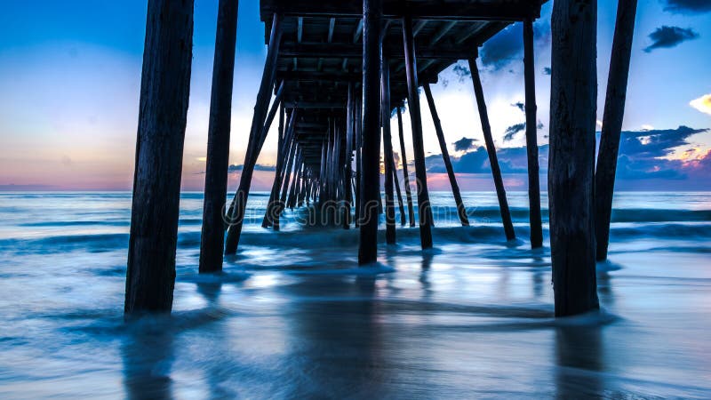 Beautiful long exposure ocean sunrise and gentle waves beneath an old wooden fishing pier. Colors of morning light in the sky. royalty free stock photo Beautiful long exposure ocean sunrise and gentle waves beneath an old wooden fishing pier. Colors of morning light in the sky. royalty free stock photo