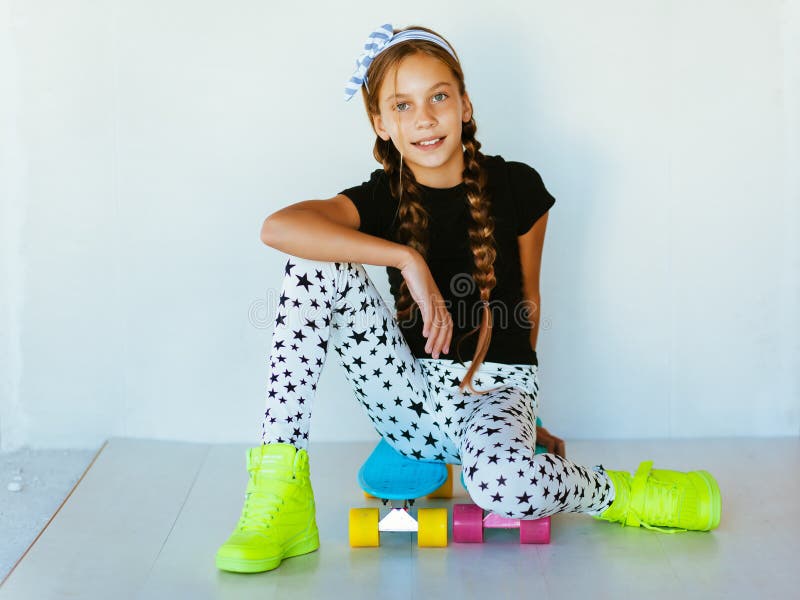 Pre teen girl wearing cool fashion clothing and sneakers posing with colorful skateboard against white wall. Pre teen girl wearing cool fashion clothing and sneakers posing with colorful skateboard against white wall