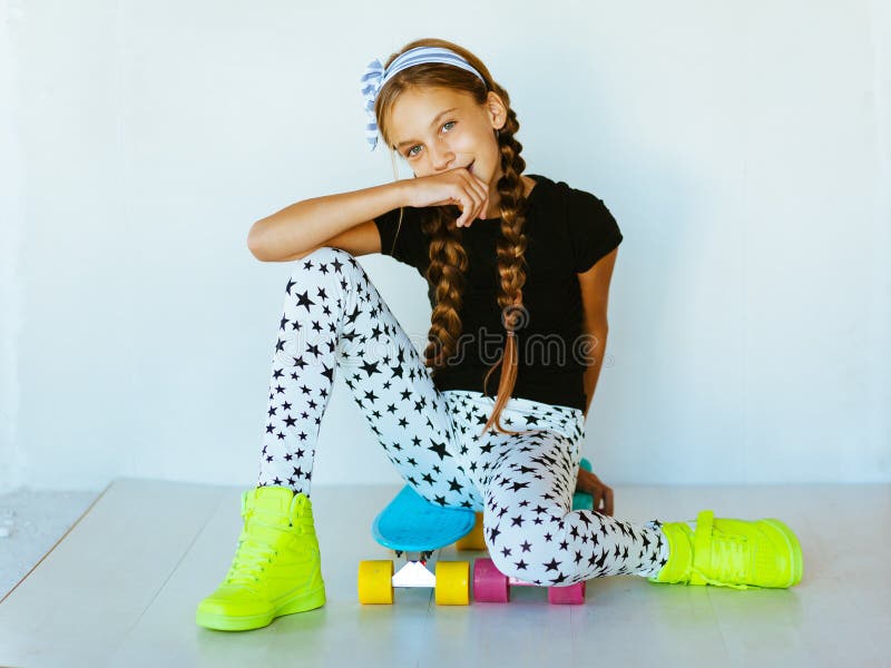 Pre teen girl wearing cool fashion clothing and sneakers posing with colorful skateboard against white wall. Pre teen girl wearing cool fashion clothing and sneakers posing with colorful skateboard against white wall