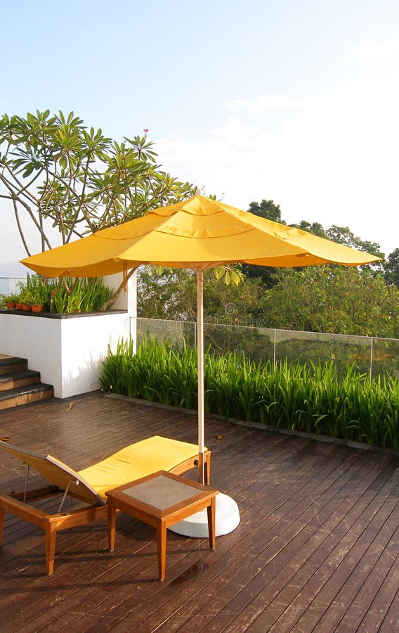 An image showing the architectural and landscaping design of a highrise garden patio of a resort hotel, furnished with simple and elegant outdoor terrace furniture - bright yellow beach sunshade with weatherproof table and lounger chairs. Landscaping with bright colorful tropical flowering trees and shrubs plants. Floor paved with weathered wooden boards. Horizontal color format. Nobody in picture. Simple composition with copy space. An image showing the architectural and landscaping design of a highrise garden patio of a resort hotel, furnished with simple and elegant outdoor terrace furniture - bright yellow beach sunshade with weatherproof table and lounger chairs. Landscaping with bright colorful tropical flowering trees and shrubs plants. Floor paved with weathered wooden boards. Horizontal color format. Nobody in picture. Simple composition with copy space.