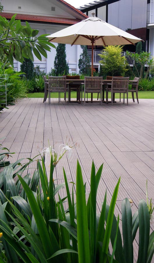 An image of a landscaped outdoors patio design, constructed with wooden decks, complete with outdoor furniture of wood table with beach sunshade shelter to suit all weather, comfortable chairs. Decorated with tropical lush landscaping all around the patio, white spider lilies flowers, frangipanni trees, junipers, lawn, shrubs etc. Upscale natural decor for landed property such as bungalow houses, villas and mansions terraces. Vertical format. Simple composition with copy space. An image of a landscaped outdoors patio design, constructed with wooden decks, complete with outdoor furniture of wood table with beach sunshade shelter to suit all weather, comfortable chairs. Decorated with tropical lush landscaping all around the patio, white spider lilies flowers, frangipanni trees, junipers, lawn, shrubs etc. Upscale natural decor for landed property such as bungalow houses, villas and mansions terraces. Vertical format. Simple composition with copy space.