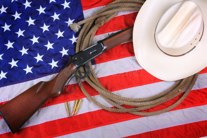 A white straw cowboy hat, a rifle with bullets and a rope laying on an American flag background. Symbol concept. A white straw cowboy hat, a rifle with bullets and a rope laying on an American flag background. Symbol concept
