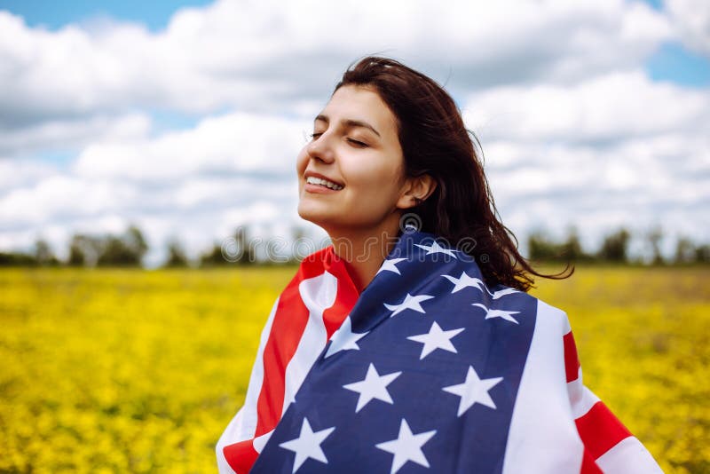 Patriotic young woman wrapped in an american flag on a beautiful field with yellow flowers. USA independence day 4th of