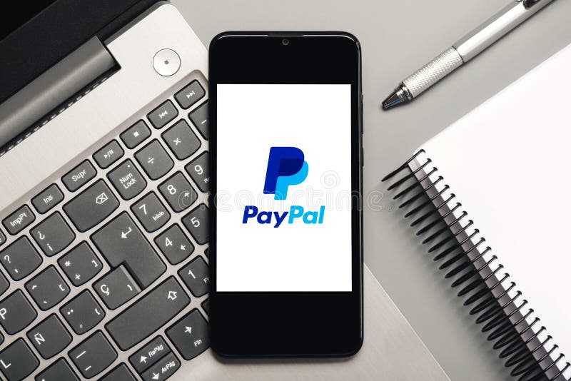 Paypal logo on black screen of smartphone with laptop and notebook. On a gray background stock image