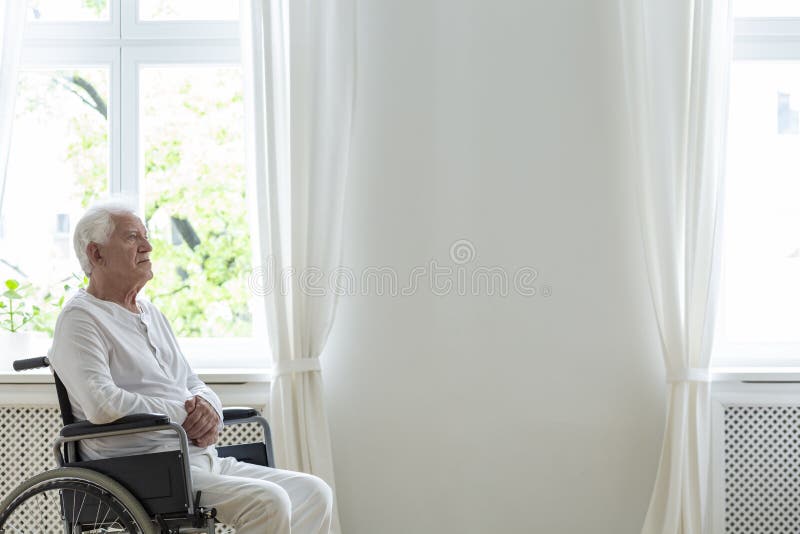 Paziente anziano solo in una sedia a rotelle in una stanza bianca accanto ad una parete vuota Disponga il vostro logo
