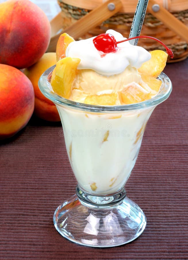 Delicious peaches stand behind a peach ice cream sundae complete with cut fresh peaches, whipped cream and a cherry.