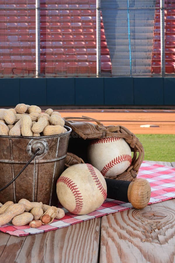 A bucket of peanuts and baseball equipment on a wood picnic table with a field and stadium in the background. A bucket of peanuts and baseball equipment on a wood picnic table with a field and stadium in the background.