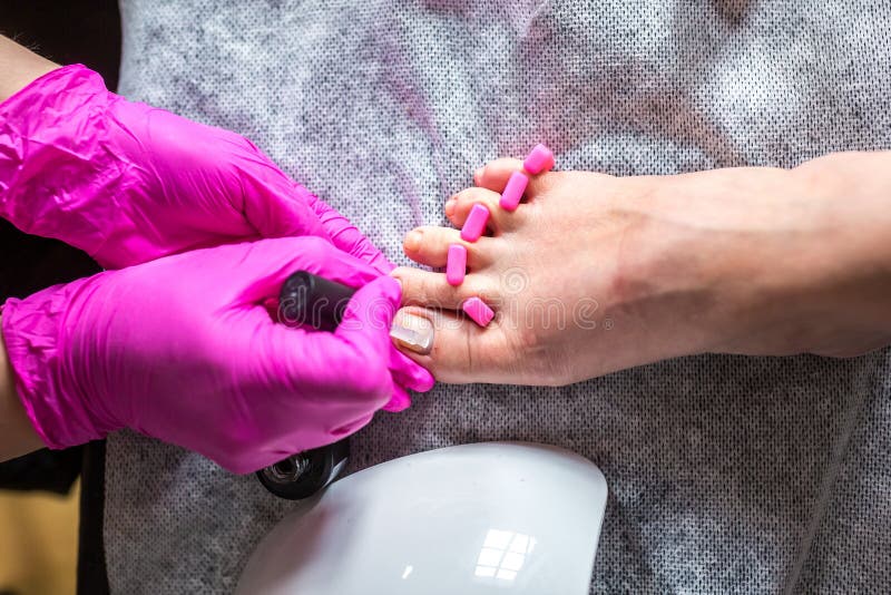 Pedicurist master in pink gloves cuts the cuticle and shellac toe nails in the pedicure salon using drill. Professional pedicure. Callous, correction.