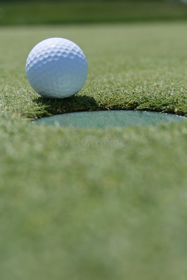 Selective focus closeup of golf ball at edge of cup against blurred background of green grass. Vertical format. Selective focus closeup of golf ball at edge of cup against blurred background of green grass. Vertical format.