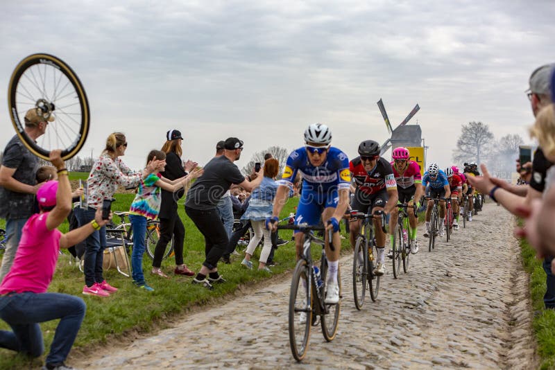 Templeuve, France - April 08, 2018: The peloton riding on the cobblestone road in Templeuve in front of the traditional Vertain Windmill during Paris-Roubaix 2018. Templeuve, France - April 08, 2018: The peloton riding on the cobblestone road in Templeuve in front of the traditional Vertain Windmill during Paris-Roubaix 2018